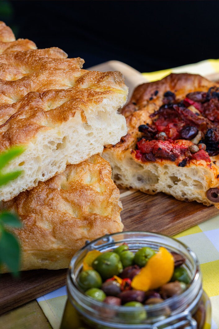 La focaccia est un classique invétéré des étés italiens. Bon, c’est vrai qu’on en mange avec grand plaisir à l’année, mais la joie de mordre dans un morceau de focaccia bien goûteuse à la plage ou lors des pique-niques al fresco assure une joie décuplée. La focaccia est un peu la cousine de la pâte pizza, et comme sa complice tomatée, elle se laisse transformer presque à l’infini. Romarin, sauge, fromage ou oignons? Oh que oui. Raisins secs, zeste d’agrumes et cannelle? Pourquoi pas! Avec cette recette de base infaillible, vous pourrez laisser votre imagination vagabonder et mettre en vedette les produits de saison qui vous font envie, comme les belles tomates cerises et les olives qu’on a choisies pour celle-ci. Un délice incontournable à ajouter à votre arsenal!